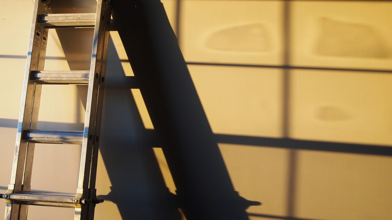 A black metal ladder leaning against a white wall in low sunlight